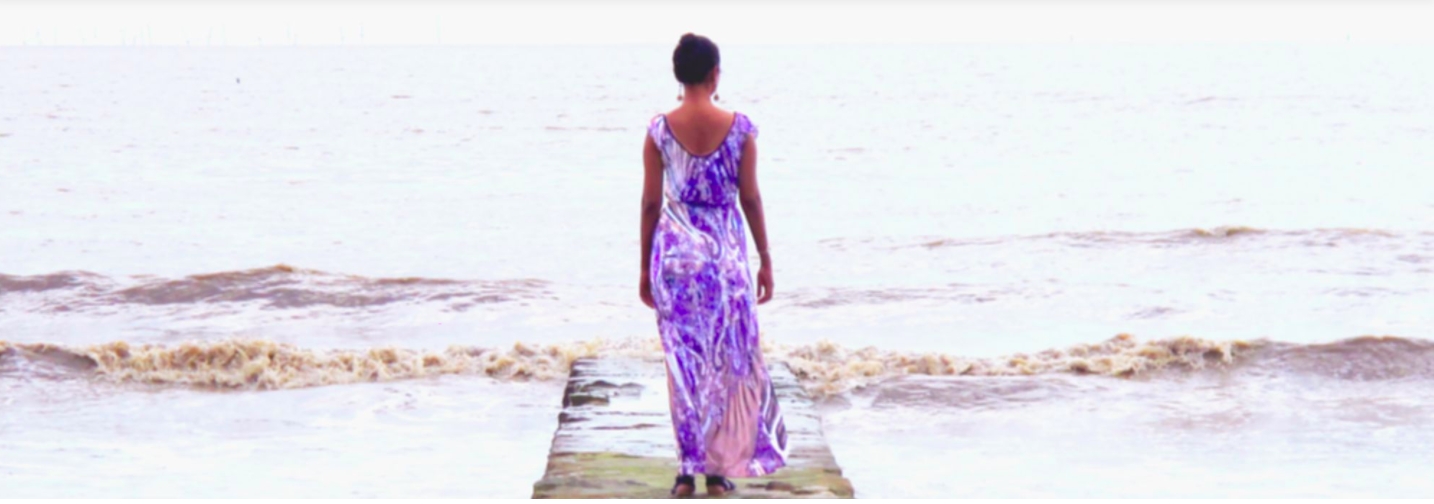 Female figure in long purple dress with hair up and wearing dangling earrings faces out towards waves